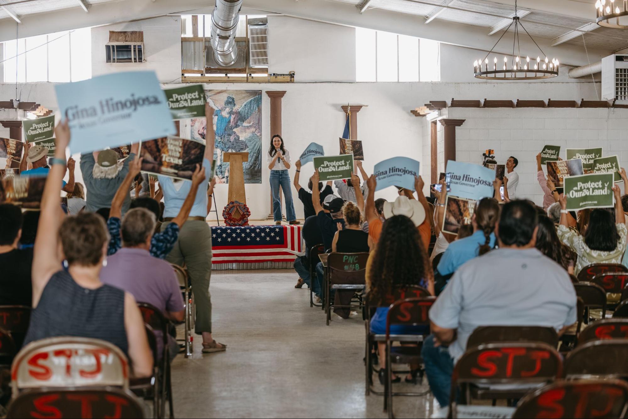 Gina speaking at an event, audience members holding "gina for texas" signs